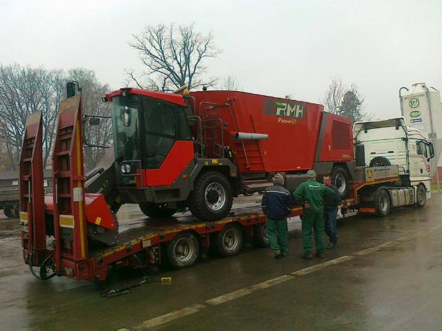 Czerwona maszyna rolnicza PMH Patron 17 transportowana na niskopodwoziowej naczepie, obok stoi biała ciężarówka, a trzech mężczyzn obserwuje załadunek.