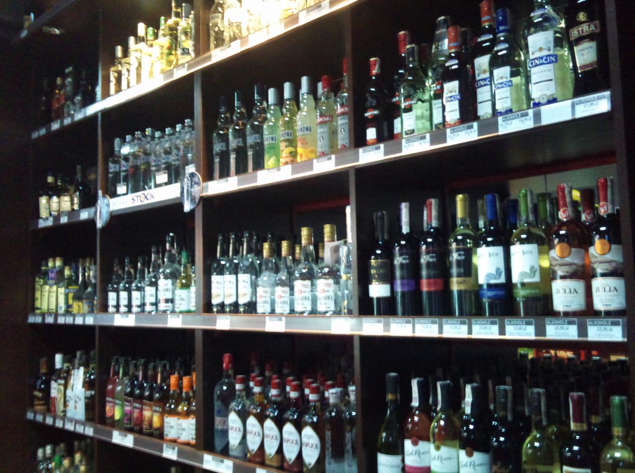Półki w sklepie z alkoholem, wypełnione różnorodnymi butelkami wódek, win i likierów. Widoczne ceny na półkach, oświetlenie punktowe.