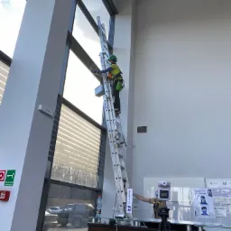 Pracownik w kasku i uprzęży, stojący na drabinie, myje wysokie okna budynku biurowego. W tle widoczny parking i inne budynki. W dolnej części kadru stanowisko do automatycznego pomiaru temperatury.