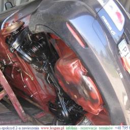 Kooperacja Polsko-Niemiecka LES-GUM Export-Import Leszek Wydrzyński - Podwozie samochodu Mazda 6 po nałożeniu czerwonej warstwy epoksydowej, czarne elementy zawieszenia, widoczny fragment tablicy rejestracyjnej i osoba w tle.