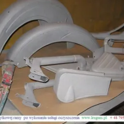 Elementy zabytkowej ramy motocykla po oczyszczeniu, przygotowane do dalszej obróbki. Widoczne błotniki, rama i inne części w szarym, matowym wykończeniu.