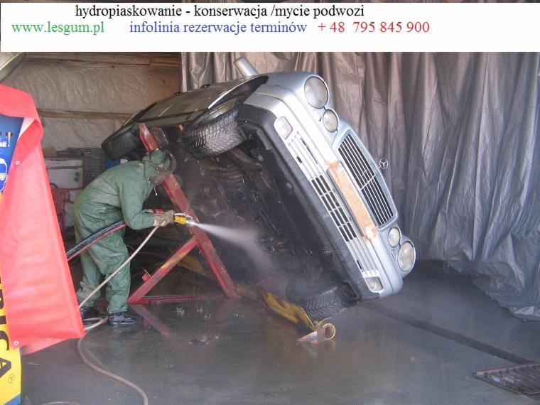Mycie podwozia srebrnego Mercedesa, umieszczonego na specjalnym stelażu, przez pracownika w kombinezonie ochronnym. Woda pod ciśnieniem.