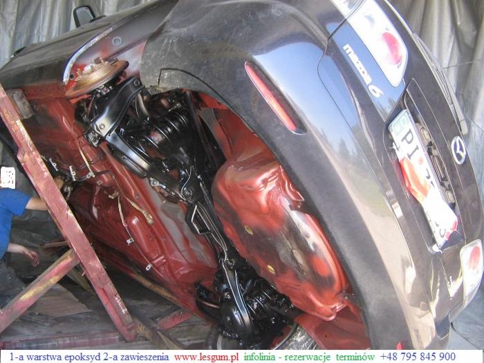 Podwozie samochodu Mazda 6 po nałożeniu czerwonej warstwy epoksydowej, czarne elementy zawieszenia, widoczny fragment tablicy rejestracyjnej i osoba w tle.
