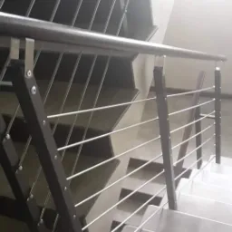 Schody z szarymi stopniami i balustradą z drewnianymi słupkami połączonymi cienkimi, poziomymi, metalowymi prętami.