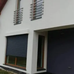 Nowoczesny dom z białymi ścianami, oknami z roletami zewnętrznymi i balkonami z poziomymi, metalowymi barierkami, widok z zewnątrz na trawnik i paletę budowlaną.