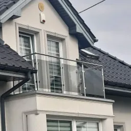 Balkon z nowoczesną szklaną balustradą z elementami ze stali nierdzewnej w jasnej elewacji domu z ciemną dachówką i wystającym gzymsem.