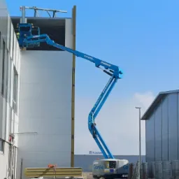 Niebieski podnośnik koszowy marki Genie, z operatorem w koszu, używany do montażu elewacji budynku przemysłowego, z widocznymi panelami elewacyjnymi i logo Husqvarna w tle.