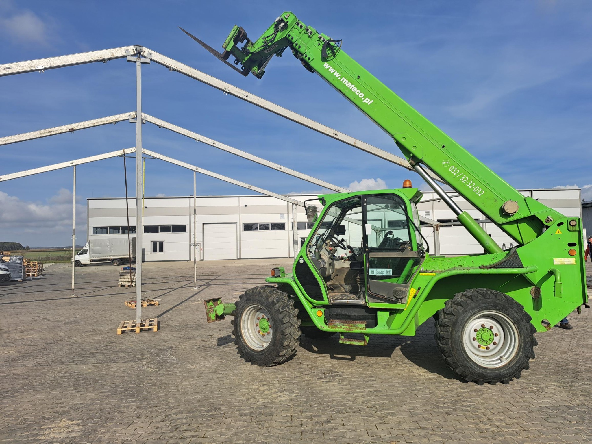 Zielony wózek widłowy teleskopowy z podniesionym ramieniem na placu budowy, w tle konstrukcja stalowa hali i elementy montażowe namiotu.