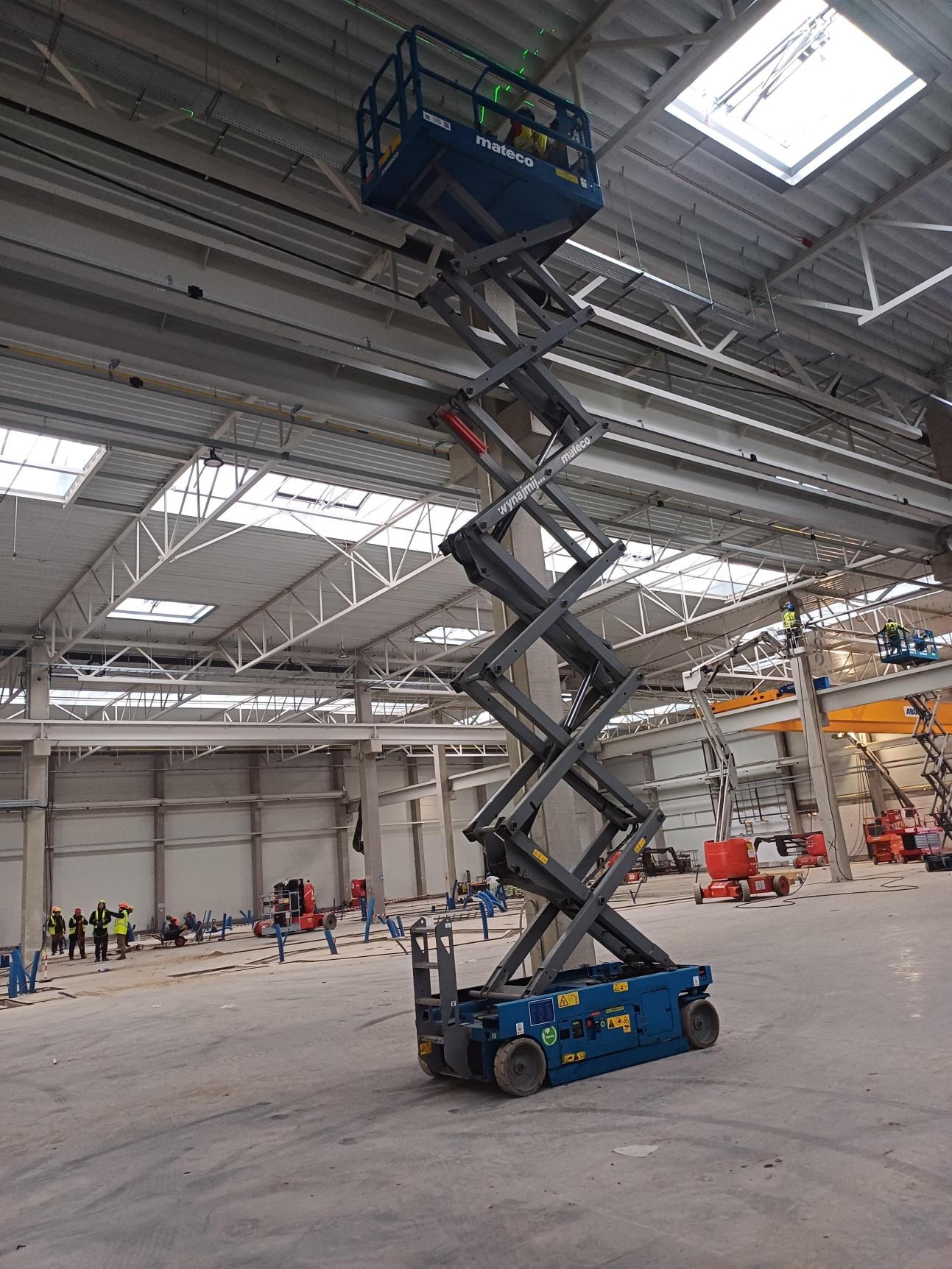 Niebieski podnośnik nożycowy Mateco w hali produkcyjnej z widocznymi pracownikami i elementami konstrukcyjnymi dachu.