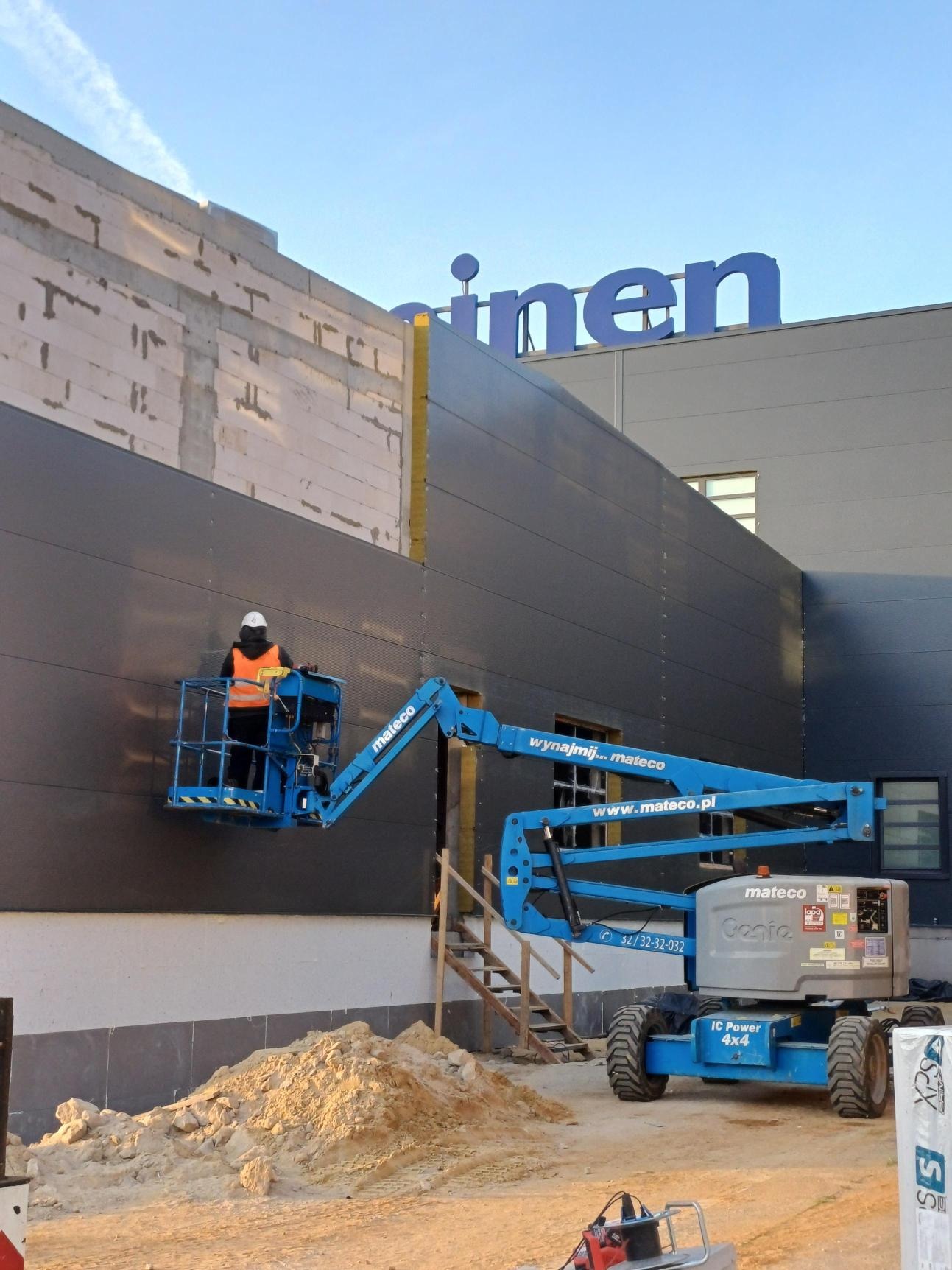 Pracownik w pomarańczowej kamizelce na niebieskim podnośniku koszowym marki Mateco, montuje elewację budynku z szarych paneli, na dachu widoczny niebieski napis 'inen'.