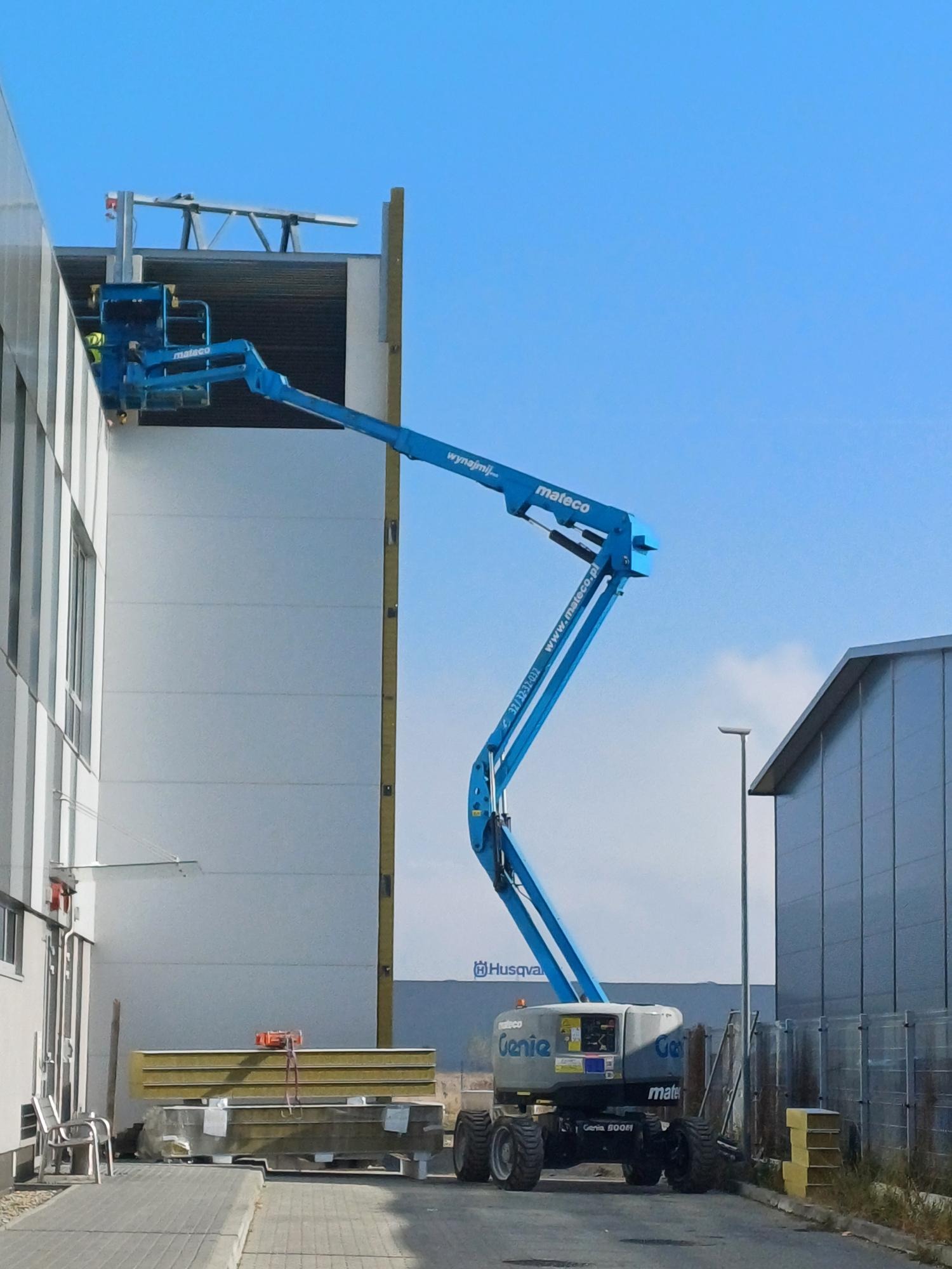 Niebieski podnośnik koszowy marki Genie, z operatorem w koszu, używany do montażu elewacji budynku przemysłowego, z widocznymi panelami elewacyjnymi i logo Husqvarna w tle.