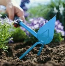 Niebieskie motyczka ogrodowa wbita w ziemię, w tle zielone i fioletowe rośliny, dłoń ogrodnika w górnym lewym rogu kadru.