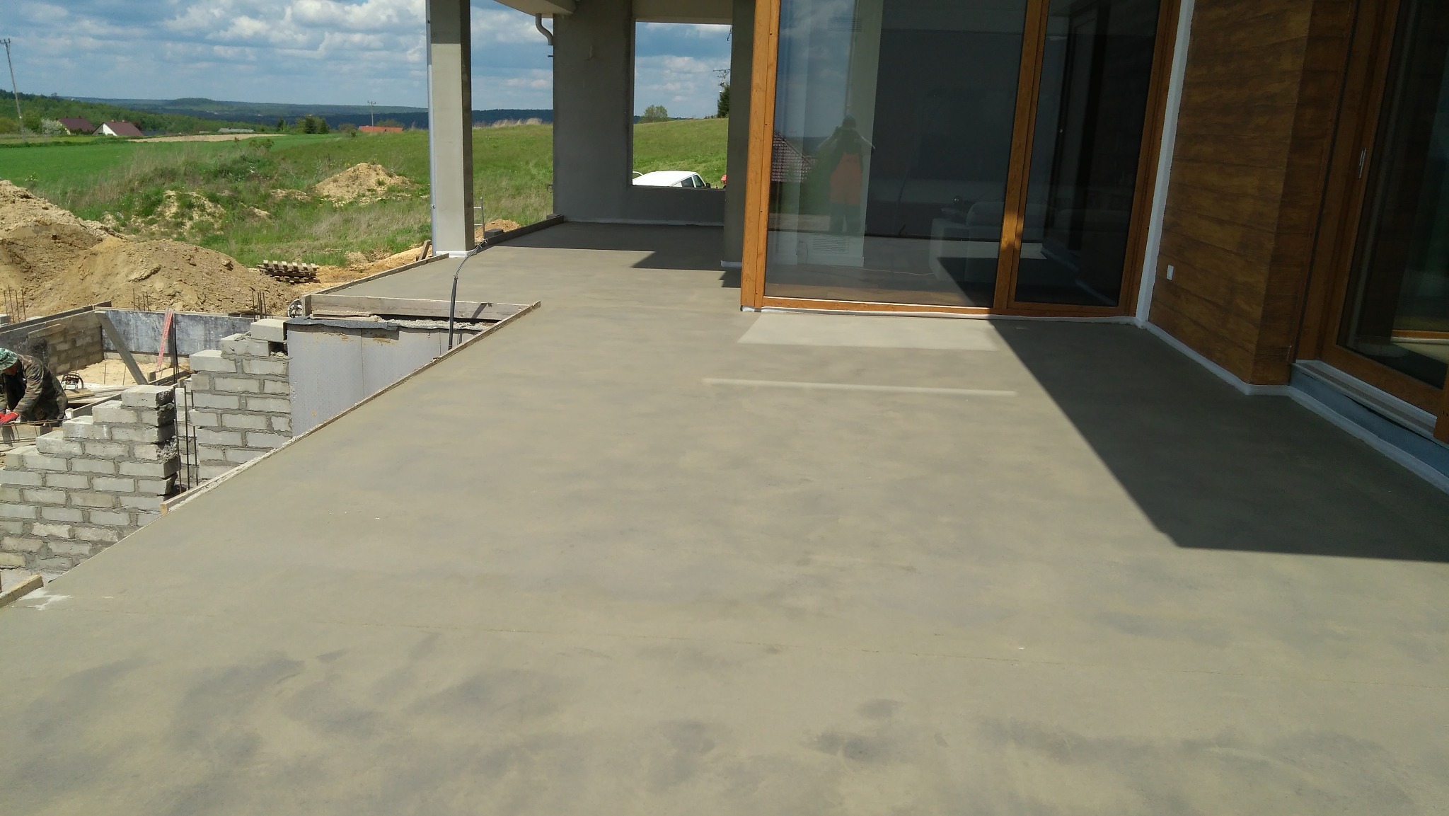 Świeżo wylana, szara posadzka jastrychowa na tarasie domu w trakcie budowy, widoczne fundamenty i krajobraz w tle.