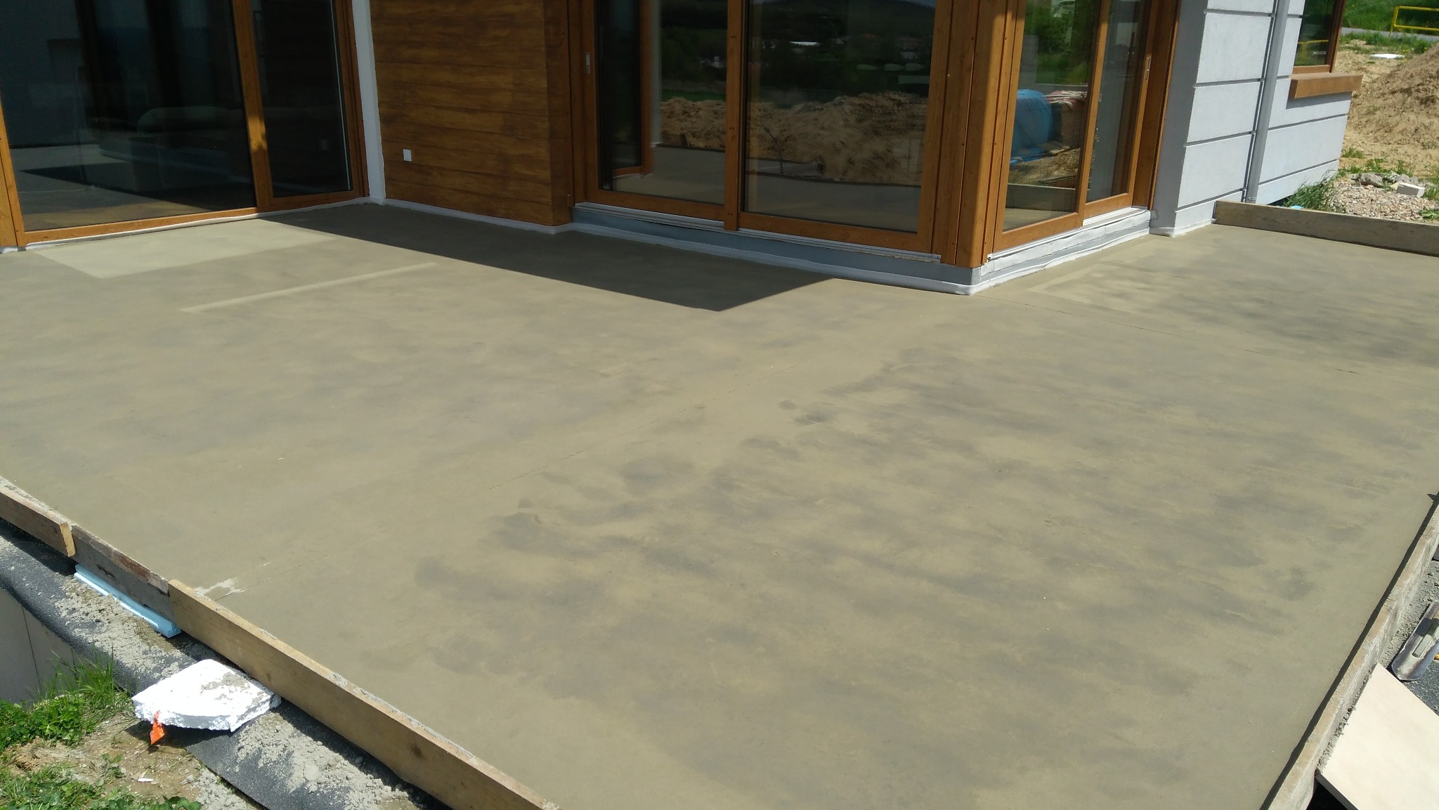Świeżo wylana, szara posadzka na zewnątrz budynku, z widocznymi drewnianymi szalunkami i styropianem izolacyjnym przy krawędziach, w tle okna tarasowe.