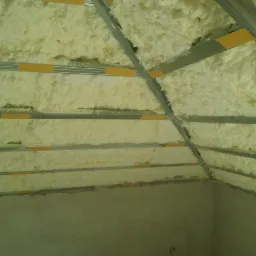 Skośny strych z widoczną izolacją piankową między metalowymi profilami, żółte taśmy mocujące, surowa ściana w dolnej części kadru.