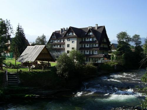 Widok na pensjonat w stylu zakopiańskim z widocznym potokiem górskim, drewnianą altaną i zielenią w otoczeniu, w tle zarys gór.