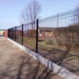 Nowoczesne, czarne panele ogrodzeniowe zamontowane na betonowej podmurówce, widok z zewnątrz posesji na tle budynku gospodarczego i drzew bez liści.