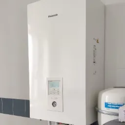 Biała jednostka wewnętrzna systemu grzewczego Panasonic Aquarea zamontowana na ścianie z białych płytek z szarym paskiem dekoracyjnym, widoczne rury i zawory, obok zbiornik wyrównawczy Airfix A.