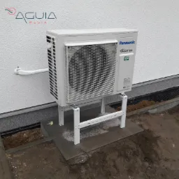 Zewnętrzna jednostka klimatyzacji Panasonic Inverter R32 umieszczona na białym, metalowym stelażu przy ścianie budynku z widocznym zabezpieczonym przewodem.