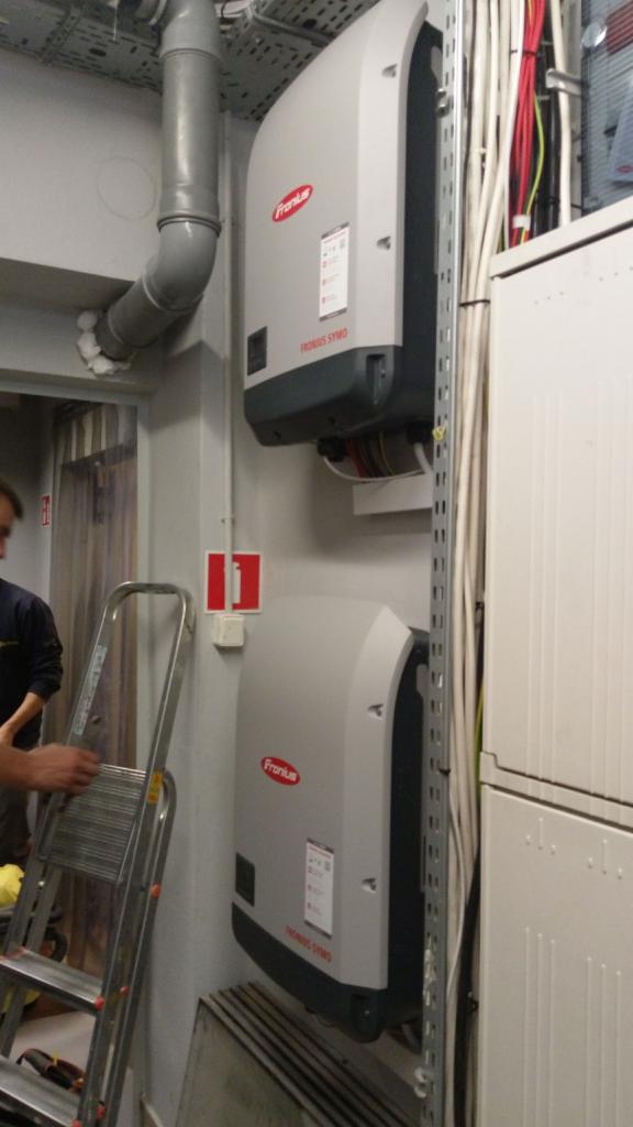 Dwa falowniki Fronius SYMO zamontowane na ścianie w pomieszczeniu technicznym, widoczne kable i rury instalacyjne, drabina w tle.