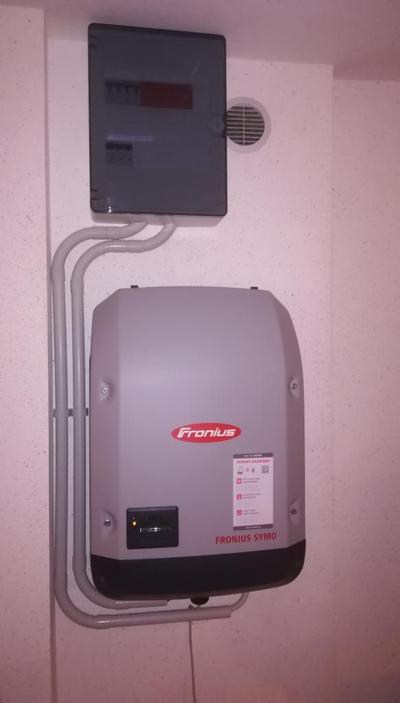 Szary falownik Fronius Symo zamontowany na ścianie z widoczną skrzynką elektryczną i wentylacją.