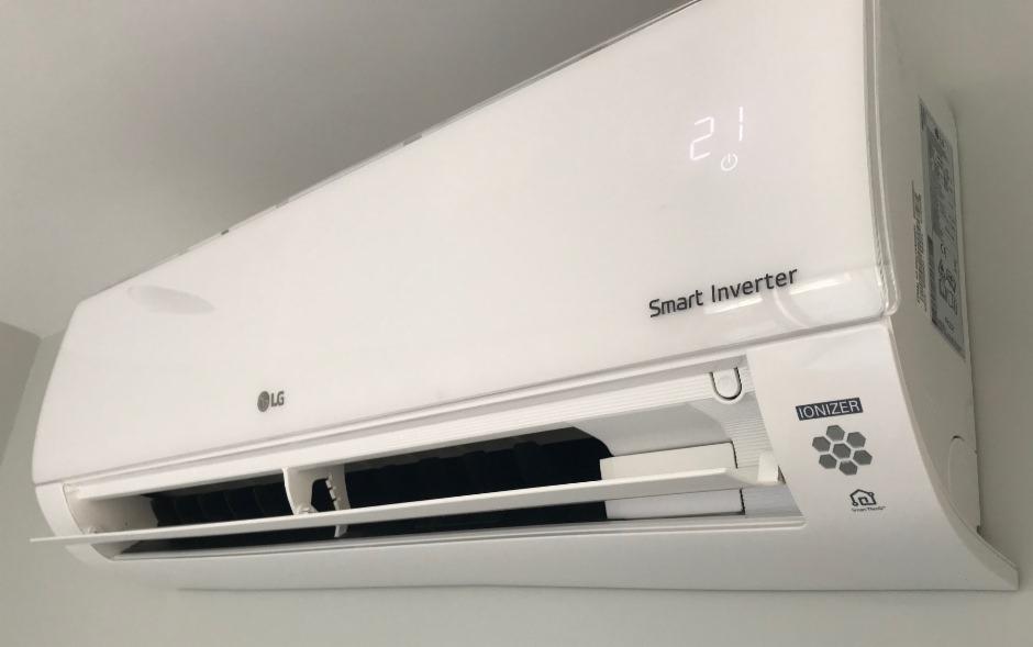 Biały klimatyzator LG Smart Inverter z otwartymi żaluzjami, wyświetlający temperaturę 21 stopni Celsjusza, zamontowany na jasnej ścianie.