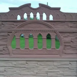 Fragment dekoracyjnego ogrodzenia z betonu w kolorze brązowym z otworami w kształcie łuków, widok na zielony trawnik za ogrodzeniem.