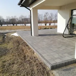 Szary taras z kostki brukowej przy nowoczesnym domu z widokiem na brzeg jeziora, obrzeża z ciemniejszej kostki, część ziemi jeszcze nie zagospodarowana.