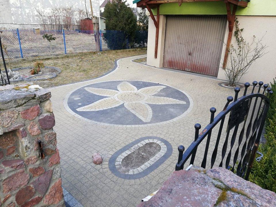 Ułożona kostka brukowa z wzorem kwiatowym na podjeździe przed garażem, widoczny fragment ogrodzenia i kamienny murek.