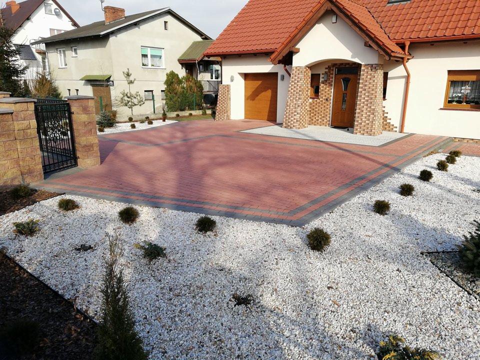 Czerwona kostka brukowa z szarymi obrzeżami tworzy podjazd do domu z garażem, otoczona białym grysem i niskimi krzewami, z widoczną bramą wjazdową z metaloplastyki.