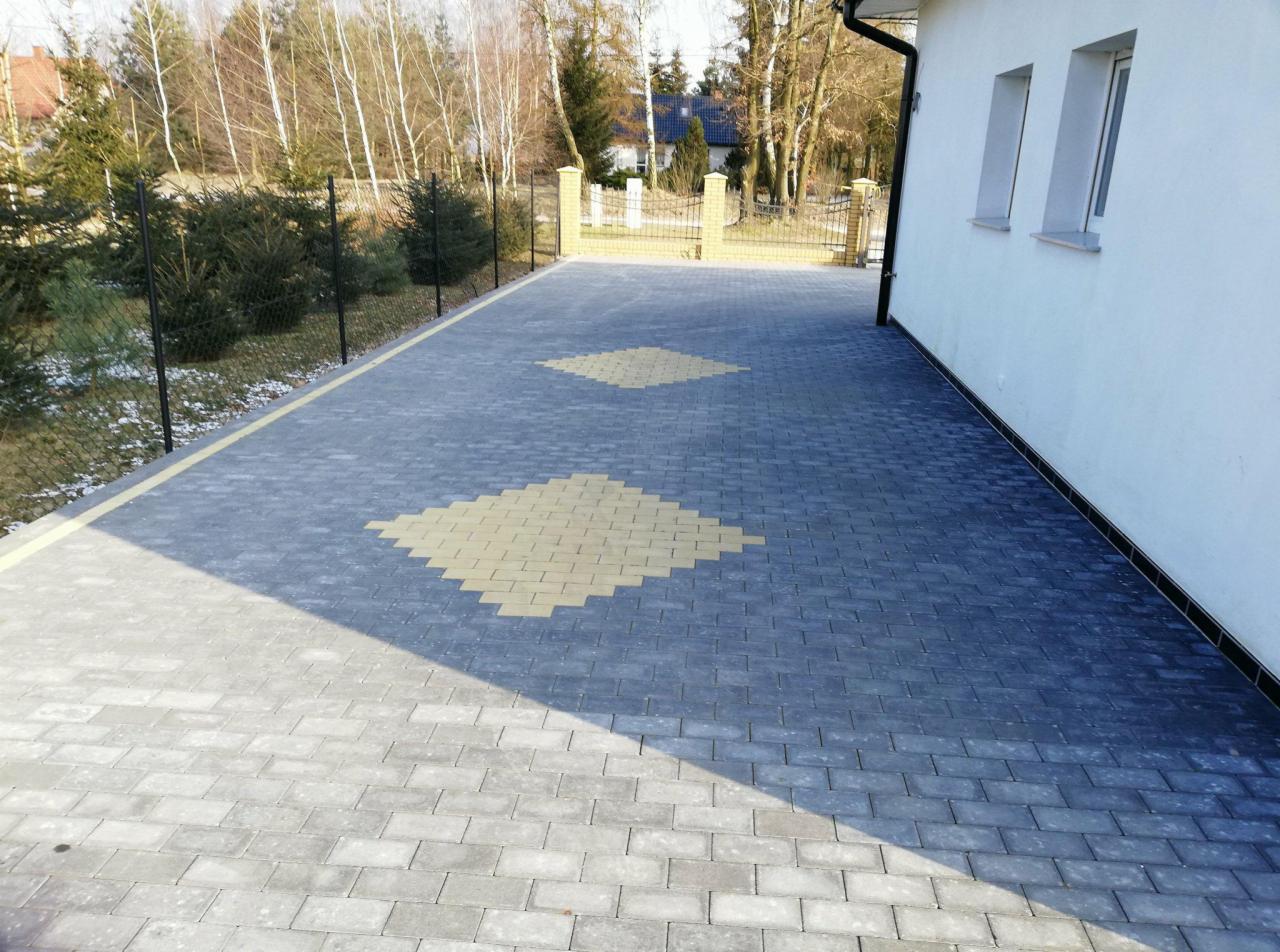 Nowoczesny podjazd z szarej kostki brukowej ułożonej we wzór z żółtymi akcentami w kształcie rombów, obok biały budynek z dwoma oknami i czarną rynną.