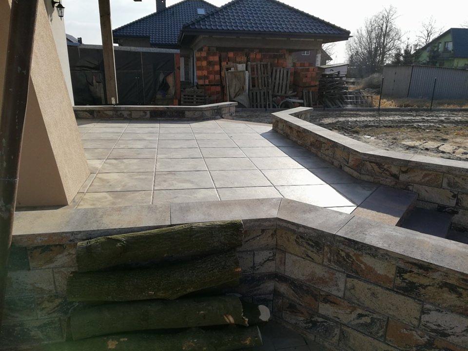 Wykończony taras z jasnych, kwadratowych płyt, obramowany murem z kamienia naturalnego o nieregularnej fakturze, z ułożonymi obok polanami drewna.