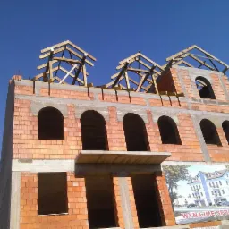 Surowy budynek z czerwonej cegły w trakcie budowy, z widoczną drewnianą konstrukcją dachu, na tle błękitnego nieba. Na dole reklama.