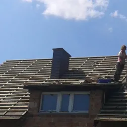 Remont dachu domu z cegły. Widoczny dekarz na dachu, komin i przygotowane łaty pod dachówki na tle błękitnego nieba.