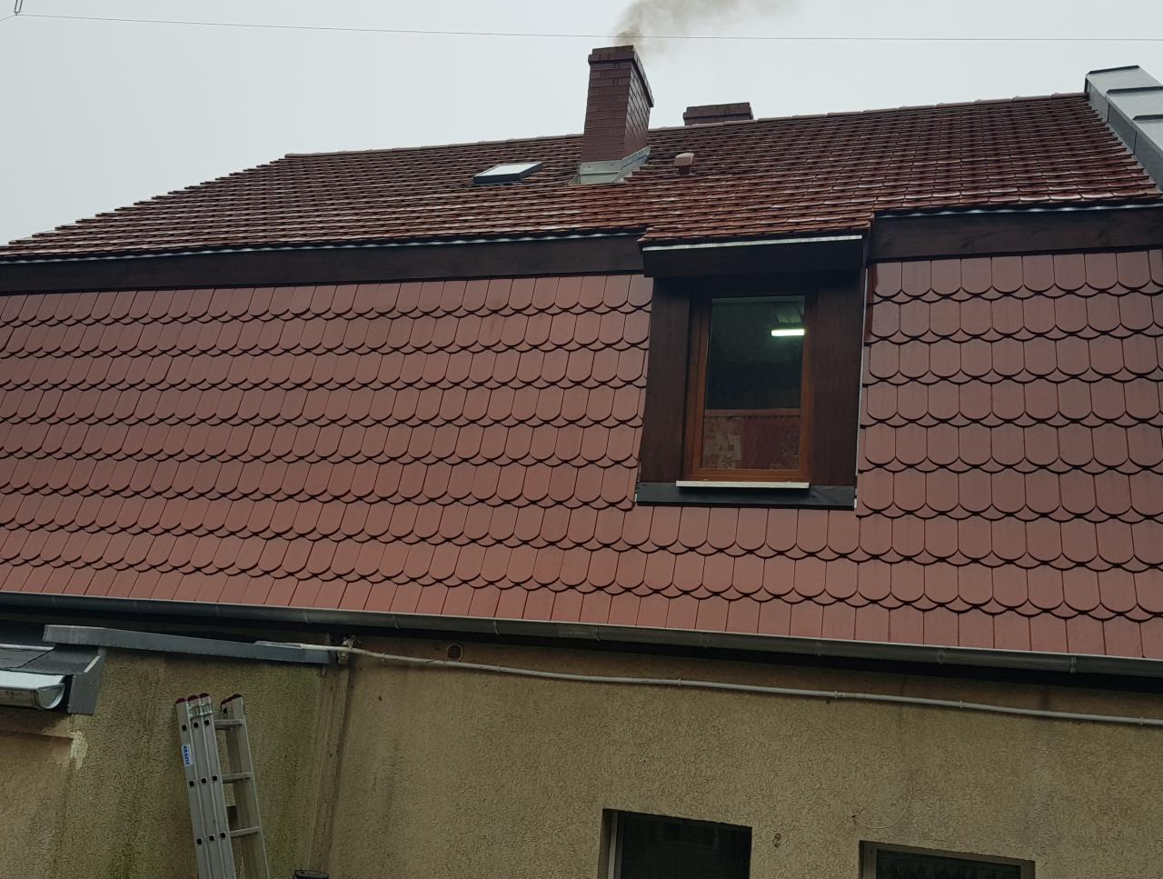Czerwona dachówka karpiówka na skośnym dachu z oknem dachowym i kominem, widoczna drabina oparta o ścianę budynku.