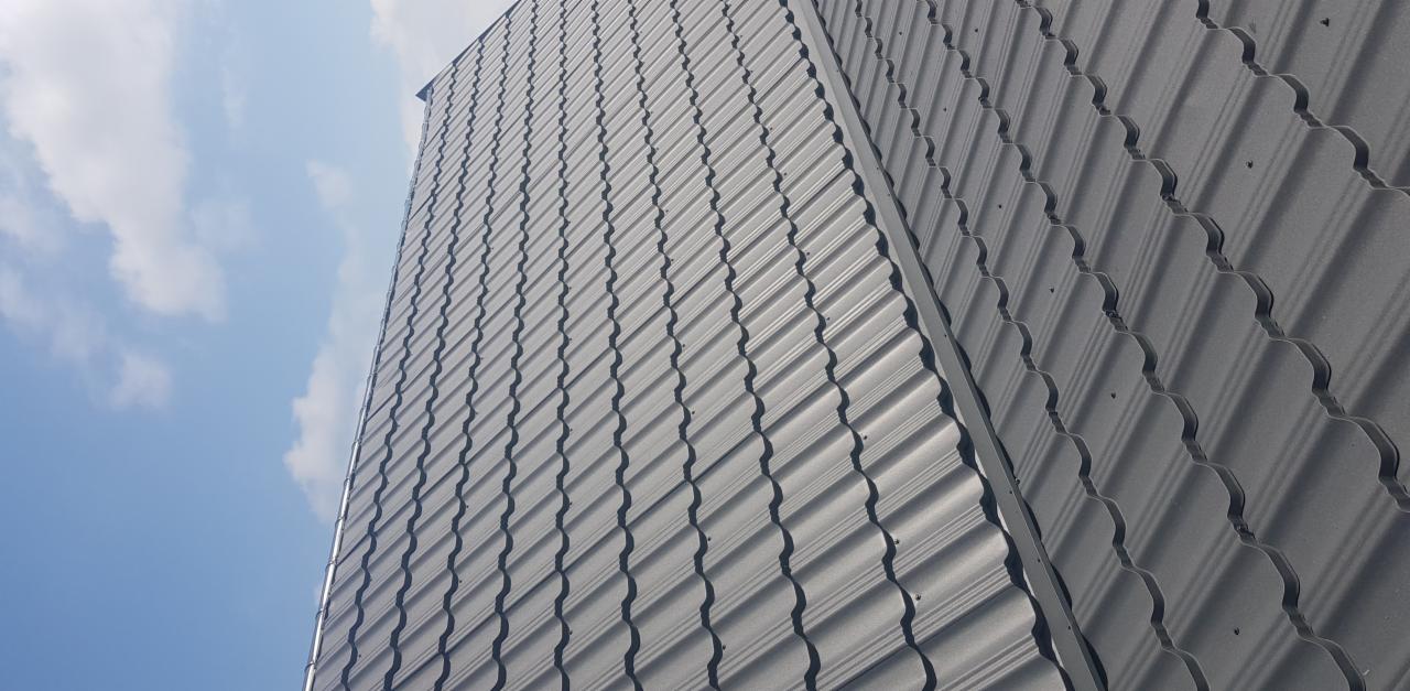 Szary dach z blachy trapezowej o falistym wzorze, widoczny z dołu pod kątem, z fragmentem błękitnego nieba z chmurami w tle.