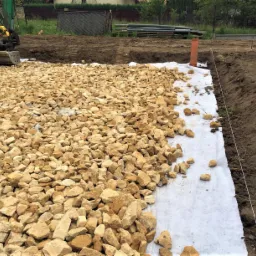 Żwirowe podłoże na białej geowłókninie, przygotowane pod budowę, z widocznym wykopem i mini koparką w tle.