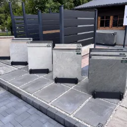 Ekspozycja różnorodnych płyt chodnikowych i paneli ogrodzeniowych na zewnątrz. Prezentacja kolorów i faktur, z detalami wykończenia i aranżacji przestrzeni.