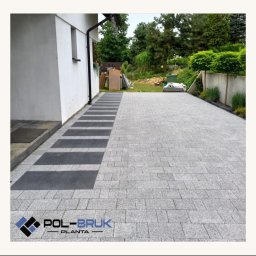 Pol-Bruk Planta Sp zoo - Jasnoszara kostka granitowa ułożona na podjeździe z ciemnoszarymi akcentami przy budynku, w tle zieleń ogrodu. Widoczny logotyp firmy.