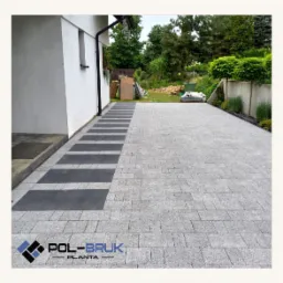 Jasnoszara kostka granitowa ułożona na podjeździe z ciemnoszarymi akcentami przy budynku, w tle zieleń ogrodu. Widoczny logotyp firmy.