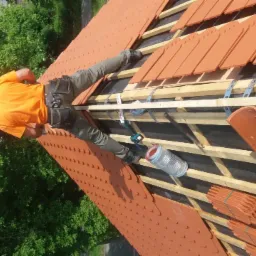 Pracownik w pomarańczowej koszulce układa czerwoną dachówkę na konstrukcji dachu, widoczne narzędzia i akcesoria dekarskie, w tle zieleń drzew.