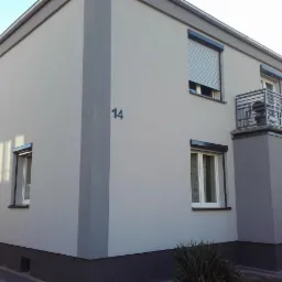 Dwukondygnacyjny dom z szarą elewacją, roletami zewnętrznymi i balkonem, numer domu 14, pracownik maluje dolną część fasady.