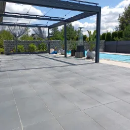 Nowoczesny taras z szarych płyt brukowych, zadaszony metalową pergolą, obok basen z niebieskim przykryciem i dmuchany różowy flaming.