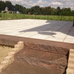 Granitowe schody prowadzą na jasny, kwadratowy taras. W tle dom i zieleń, błękitne niebo z chmurami. Krawędź tarasu z ciemnego kamienia. Widok na ogród.