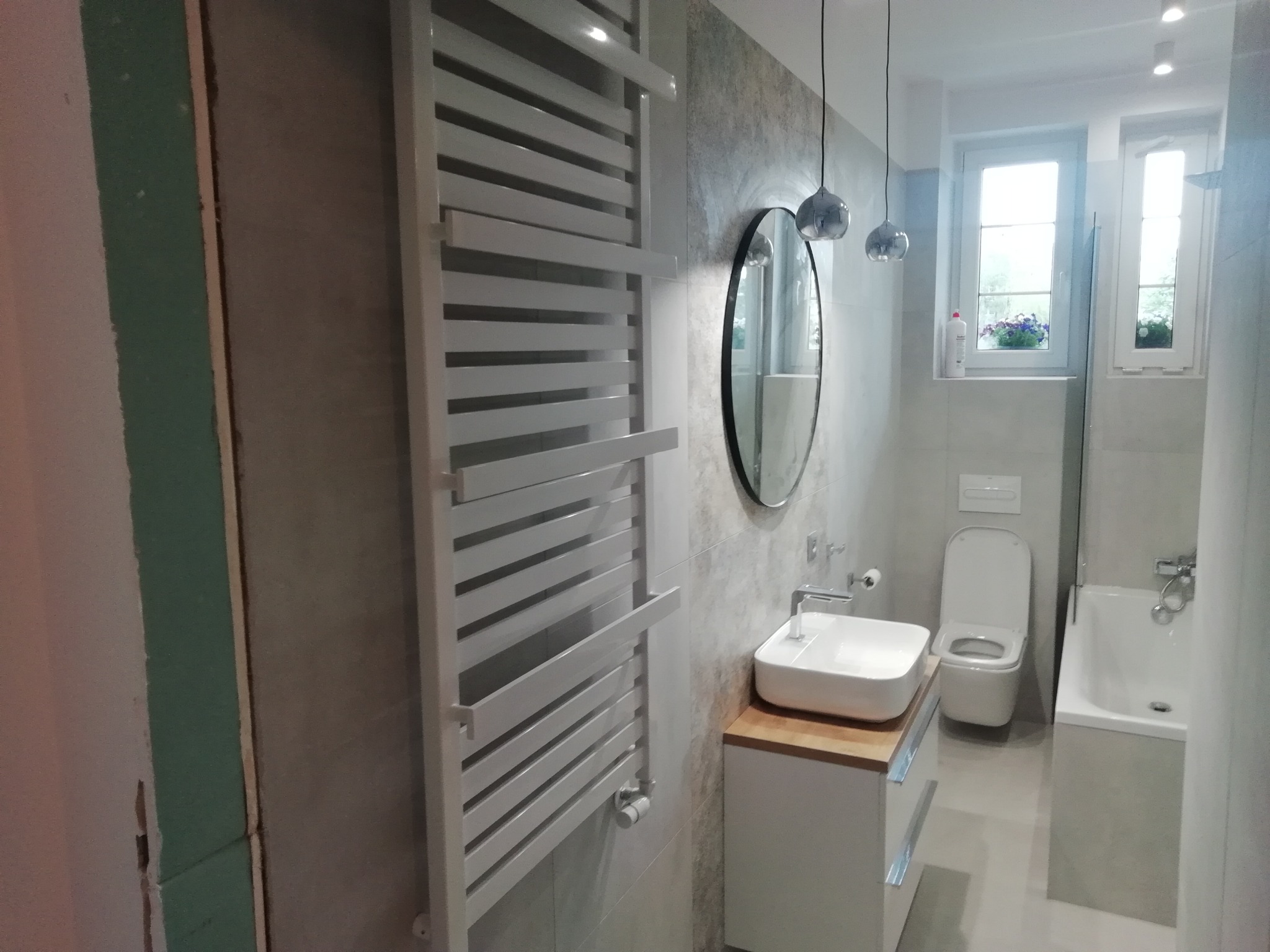 Nowoczesna łazienka w trakcie wykańczania: biały grzejnik drabinkowy, owalne lustro, umywalka nablatowa na drewnianym blacie, toaleta i wanna, ściany częściowo wykończone płytami gipsowo-kartonowymi.