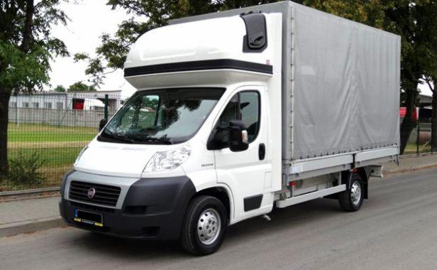 Biały bus dostawczy z plandeką na pace, zaparkowany na ulicy. Widok z boku, na tle drzew i budynków. Charakterystyczna zabudowa nad kabiną kierowcy.