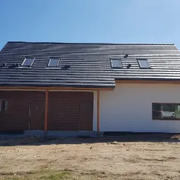 Nowoczesny dom z dwuspadowym dachem pokrytym ciemną dachówką, z czterema oknami dachowymi, częściowo wykończony drewnem, na działce w trakcie zagospodarowywania.