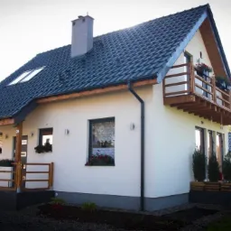 Nowoczesny dom jednorodzinny z balkonem i ciemną dachówką, widok z zewnątrz w słoneczny dzień.