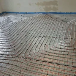 Białe rury systemu ogrzewania podłogowego ułożone spiralnie na folii izolacyjnej z czerwoną siatką, widok z góry na surowy stan wykończenia wnętrza.