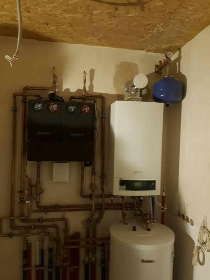 Kompletna instalacja grzewcza z kotłem, zasobnikiem i rozdzielaczem, rury miedziane, manometry, ściany w beżowym odcieniu, sufit z płyty OSB.